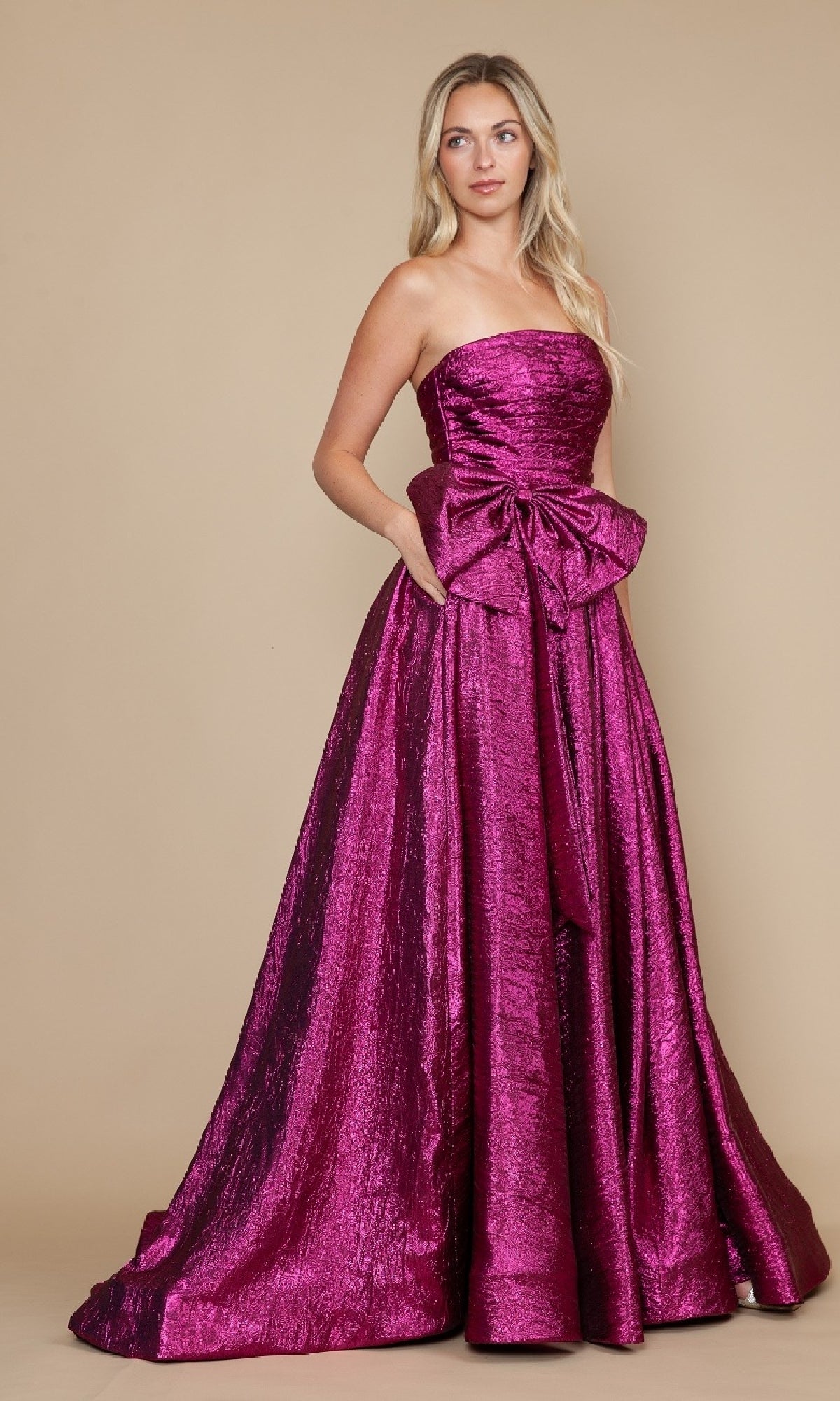 A woman wears the Poly USA 9720 Side-Bow Strapless Prom Gown in magenta, featuring an A-line silhouette and a large front bow, standing against a plain beige background.